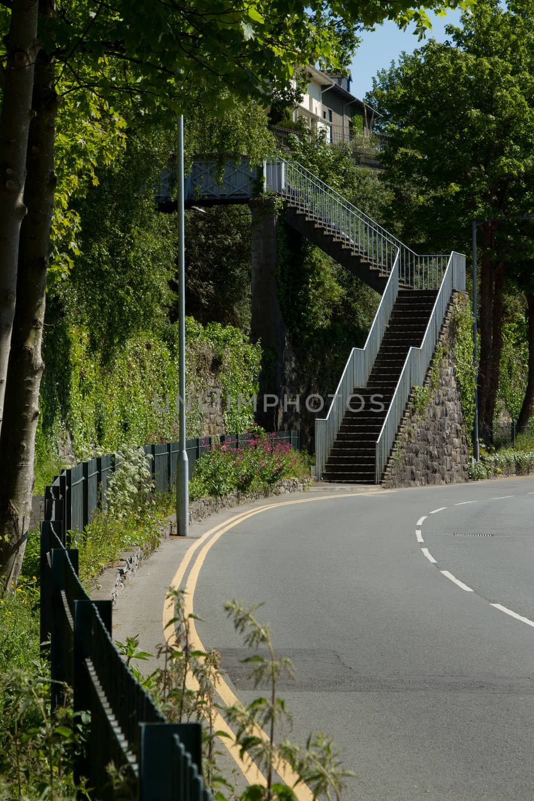 A tarmac road and fence leads to a metal set of steps leading to a footbridge.