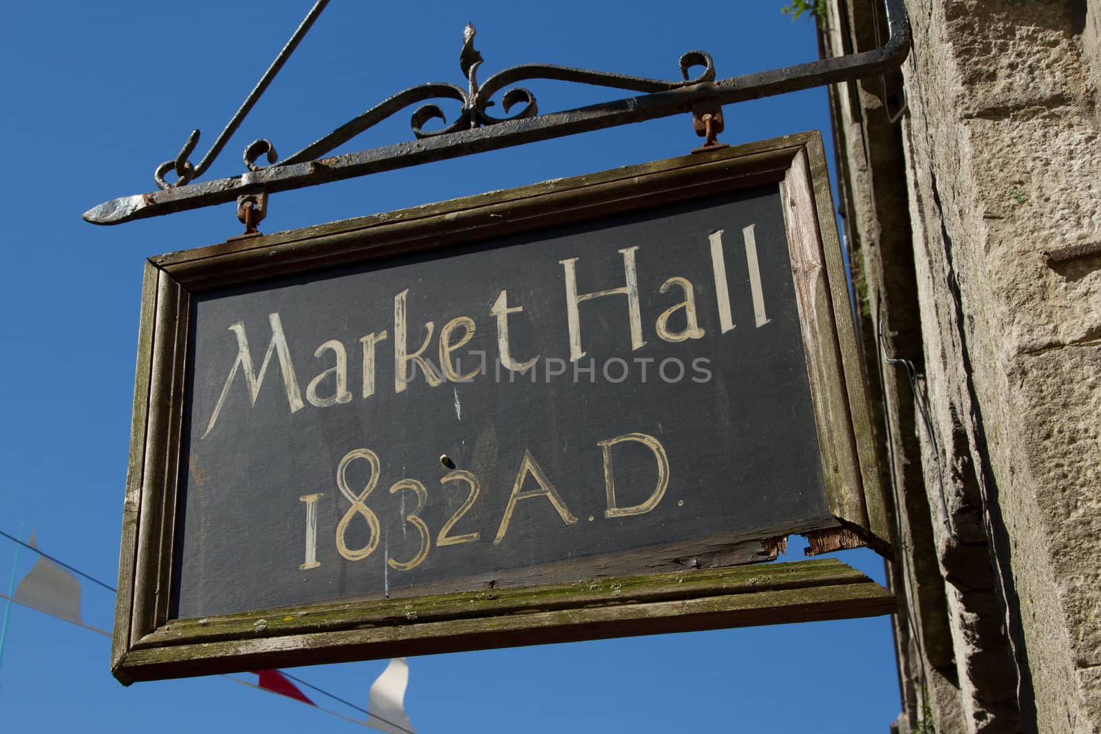 An old sign with a black board with the words 'MARKET HALL 1832 A.D.' hanging on a metal support by a broken wooden frame against a blue sky.