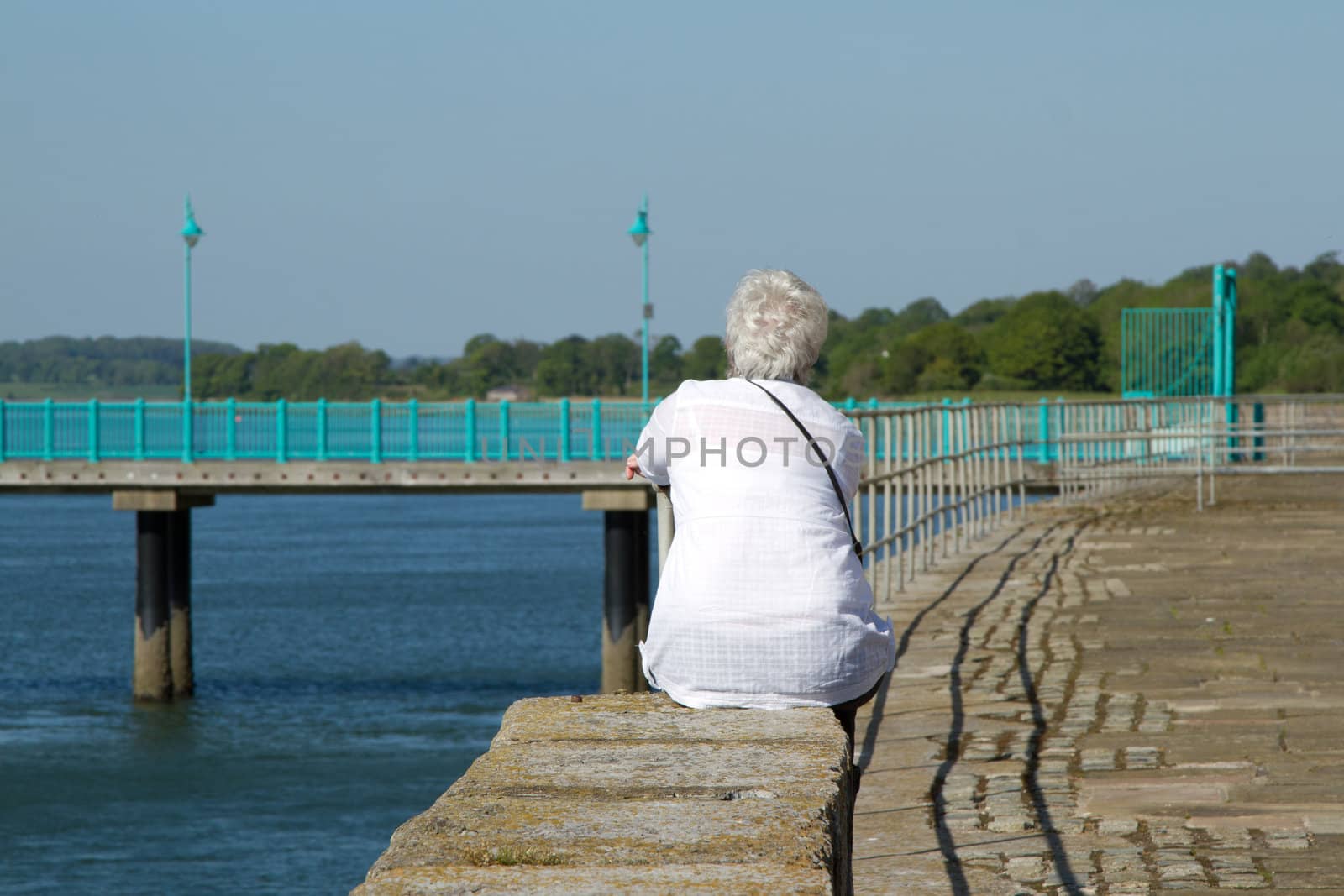 A middle aged, elder woman with short white hair, a white blouse and shoulder strap sits on a wall staring into the distance next to the sea.