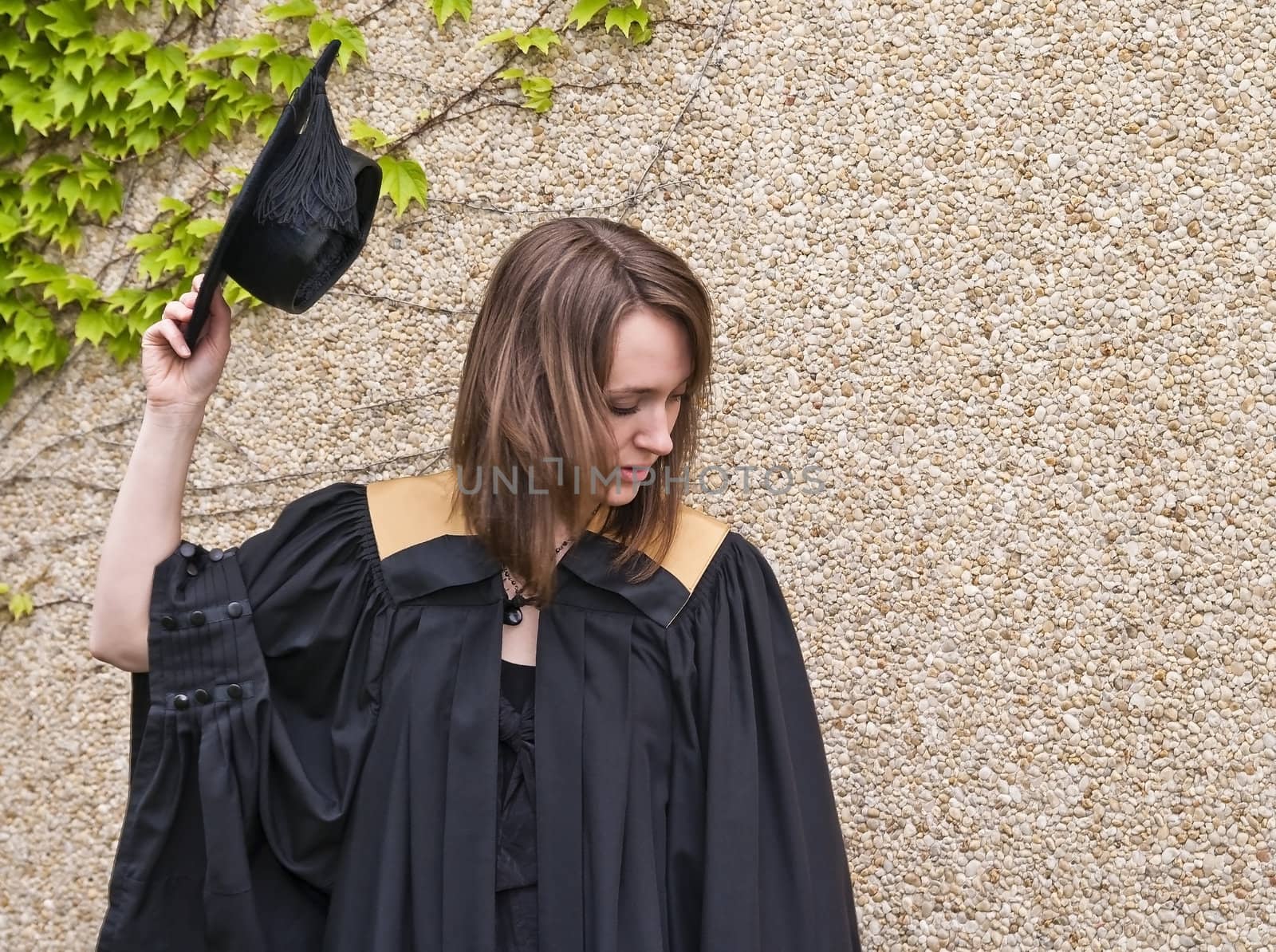 Young woman holding graduation hat against stone and leafy wall.
