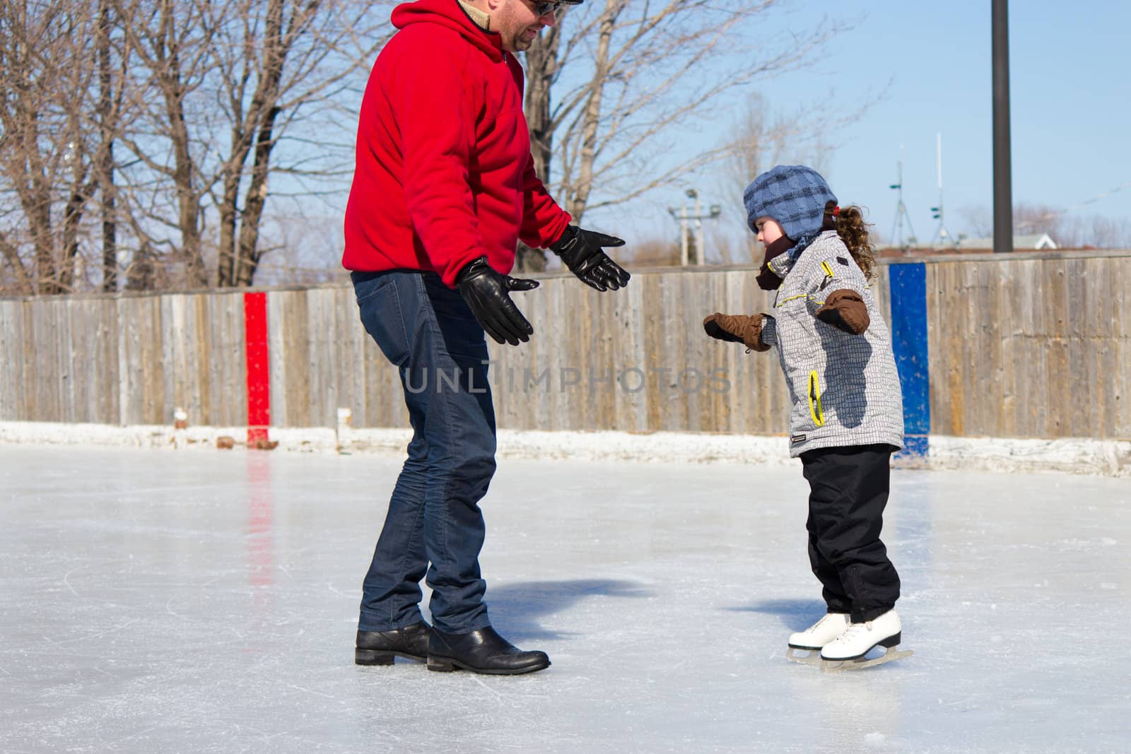 Father teaching daughter how to ice skate at an outdoor skating rink in winter.