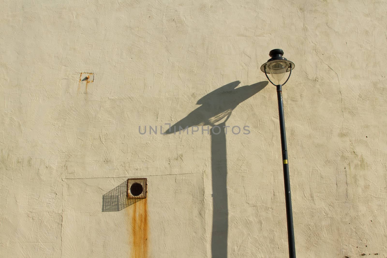 A cream painted wall with a rusty bolt and grill, rust stains and a light post with shadow.