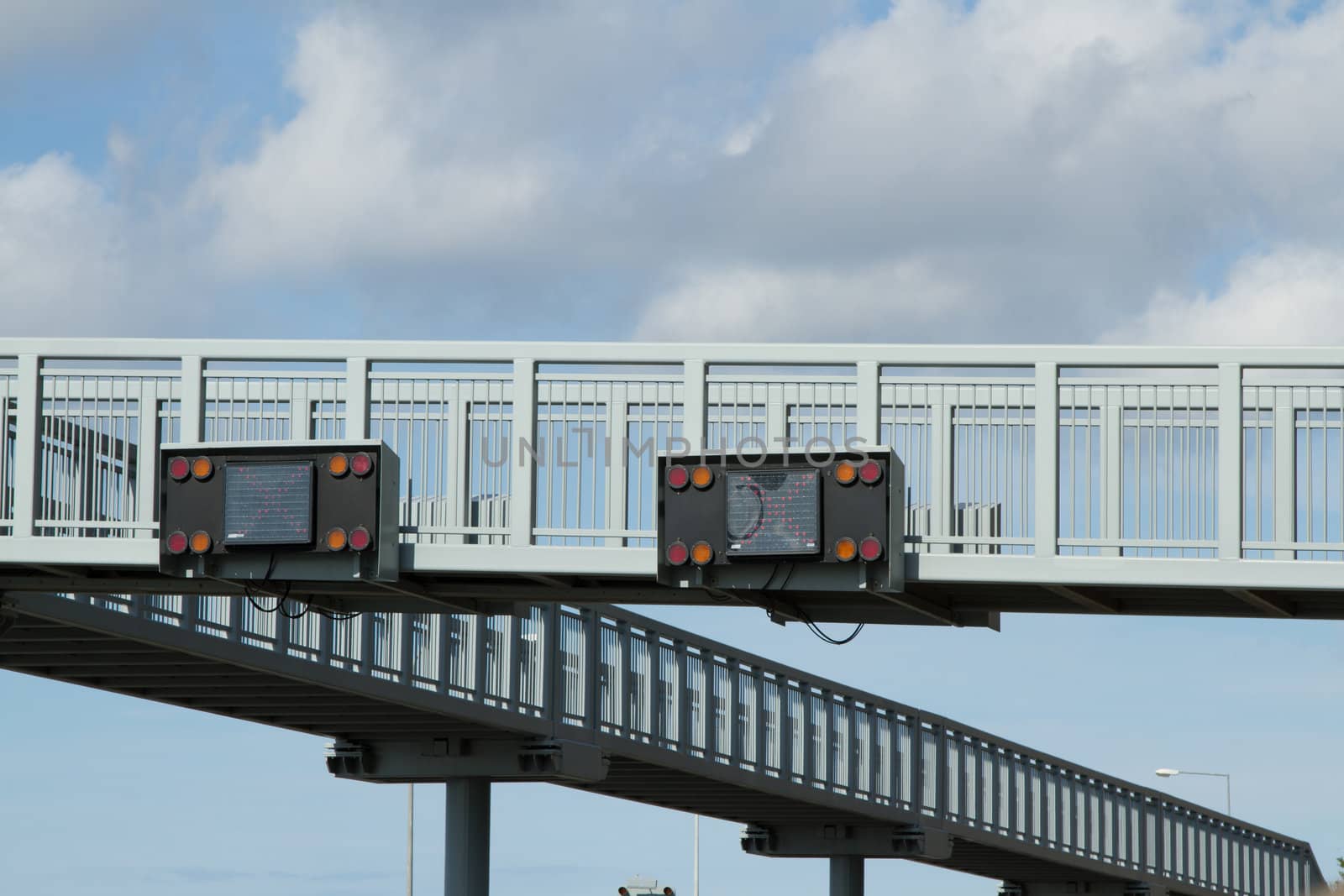 A pair of electronic traffic signs fixed to a raised metal walkway with a cloudy sky in the background.