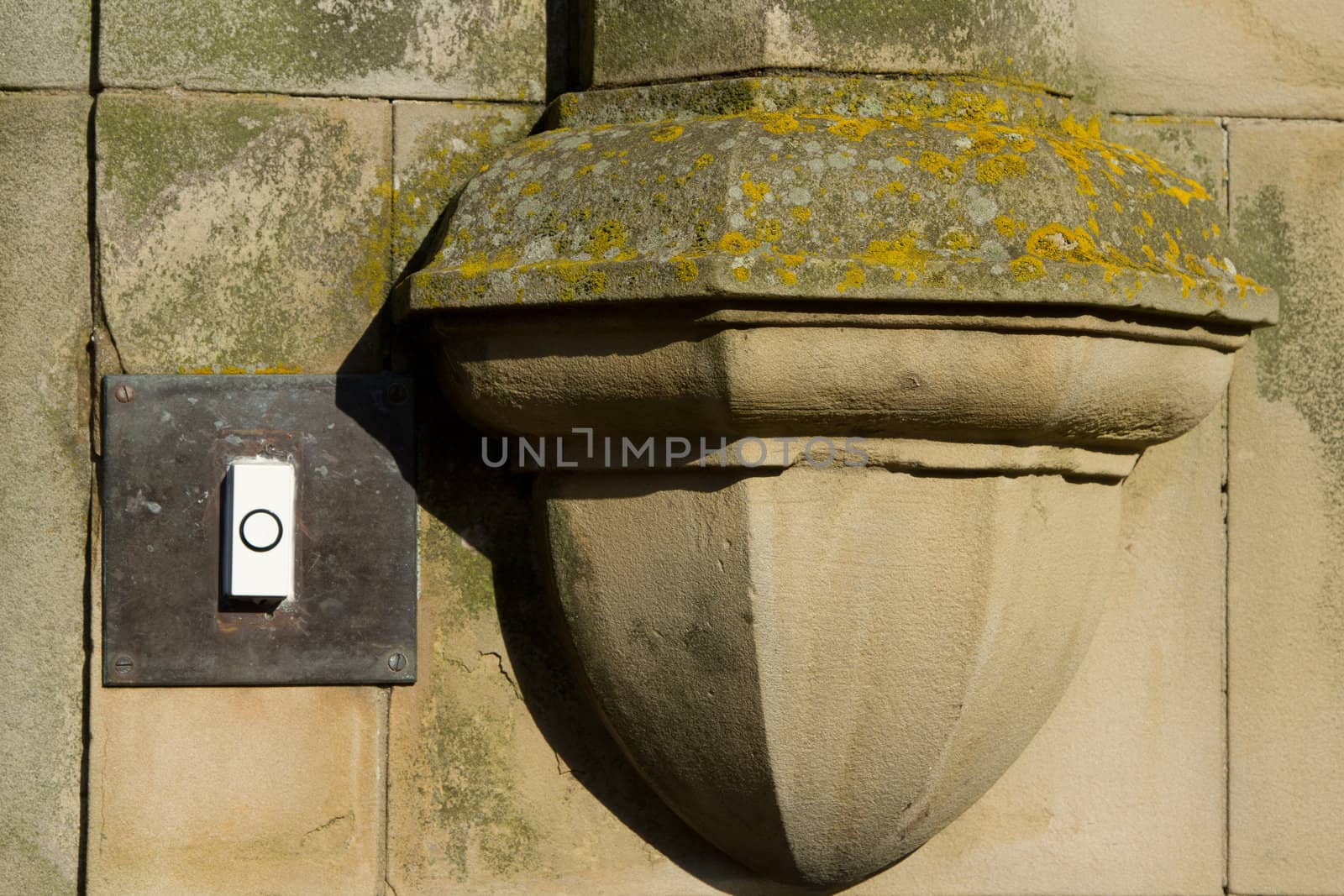 An old brass bell plate with modern white plastic bell push next to an old hand carved pillar foot on a wall with lichen and mould.