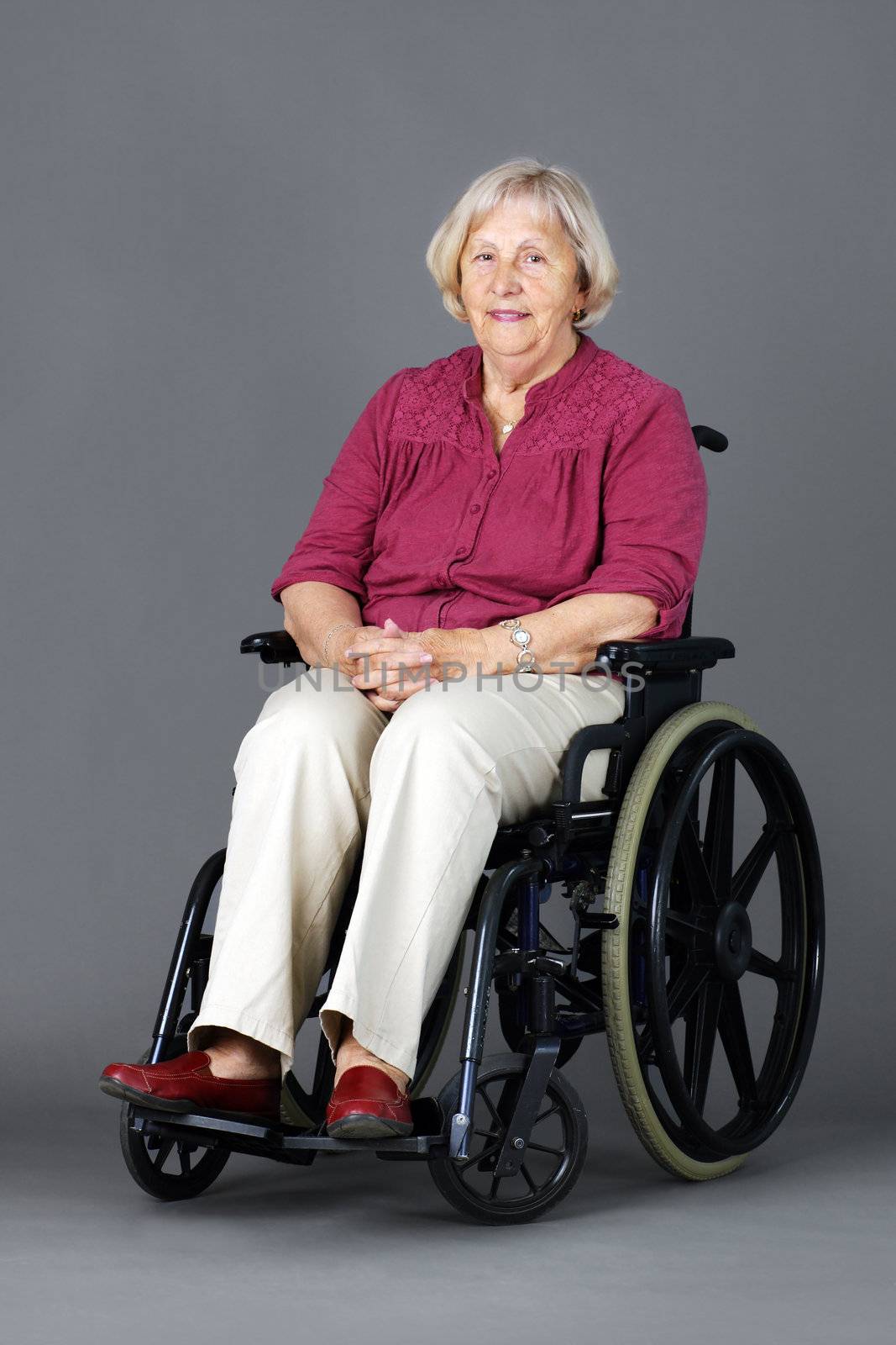 Smiling senior woman seated in a wheelchair, either handicaped or disabled, looking at camera over neutral grey background.