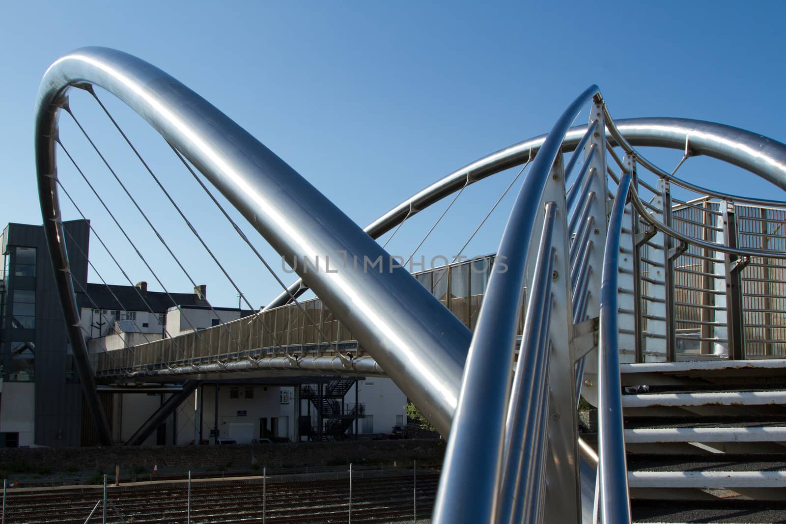 A bridge made from tubular stainless steel with steps and hand rails leading to buildings with a blue sky in the distance.