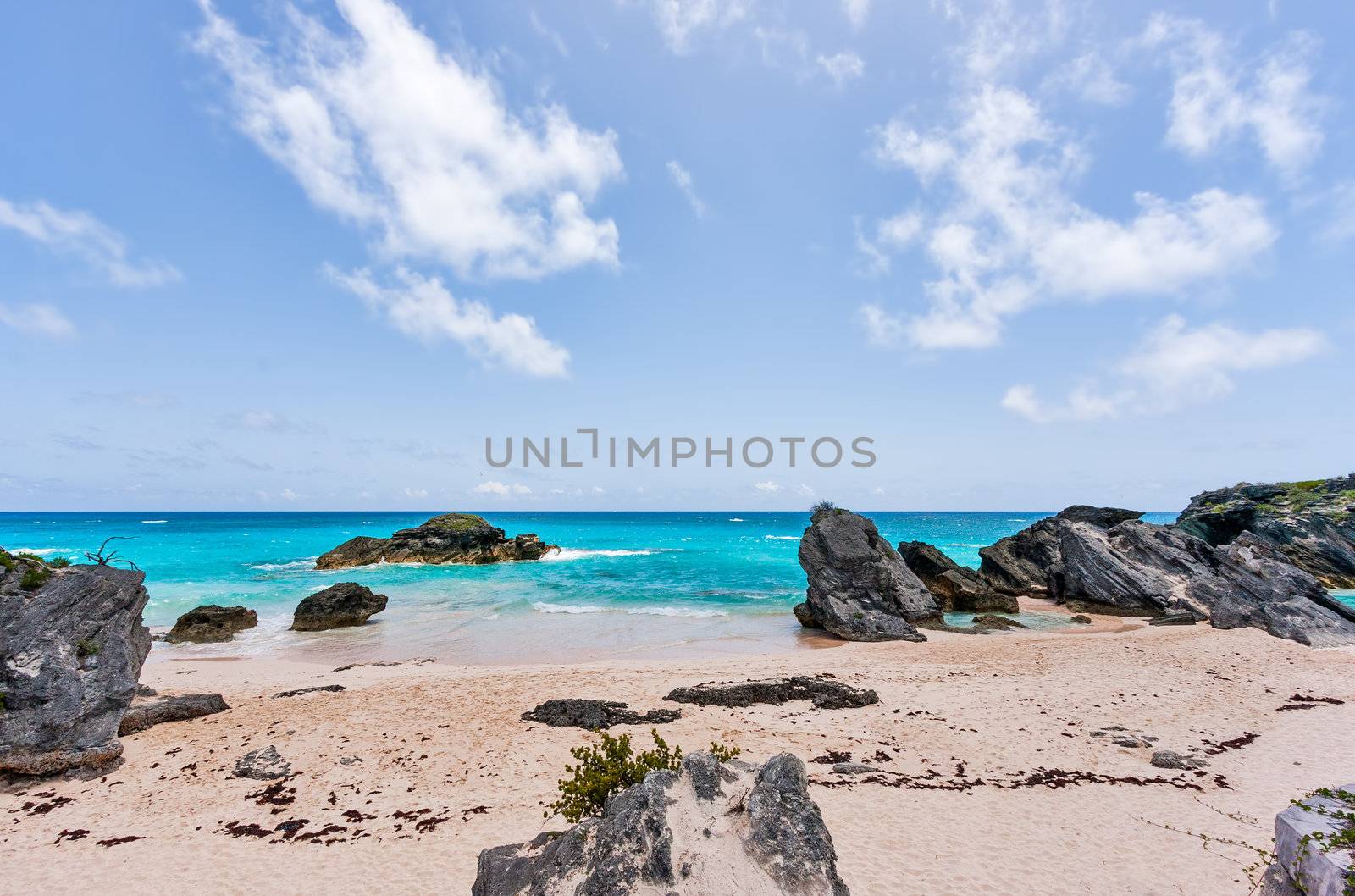 Horseshoe Bay in Bermuda. Sand and boulders are in the foreground in front of the blue water. It is a sunny day.