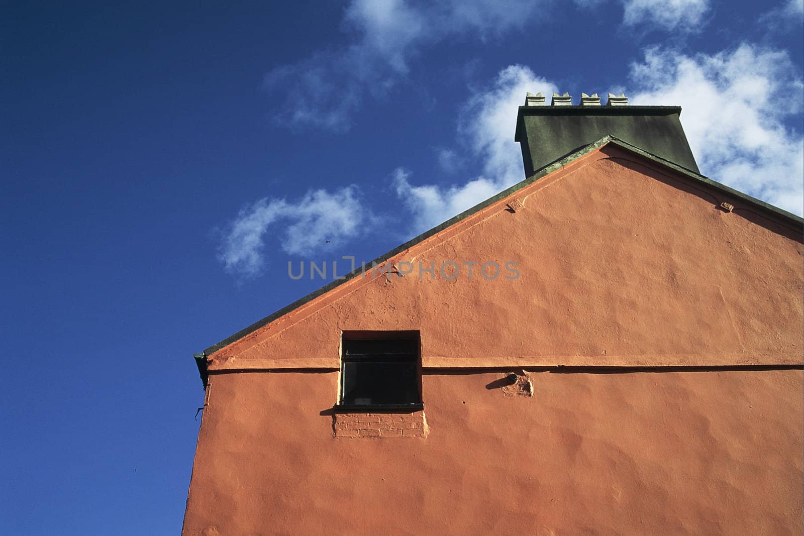 A gable end of a building painted orange with a window and chimney against a blue sky with clouds.