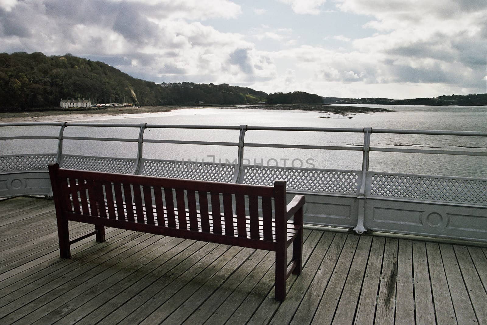 A wooden brown stained bench on boards with a railing and the sea, trees and sky in the distance.