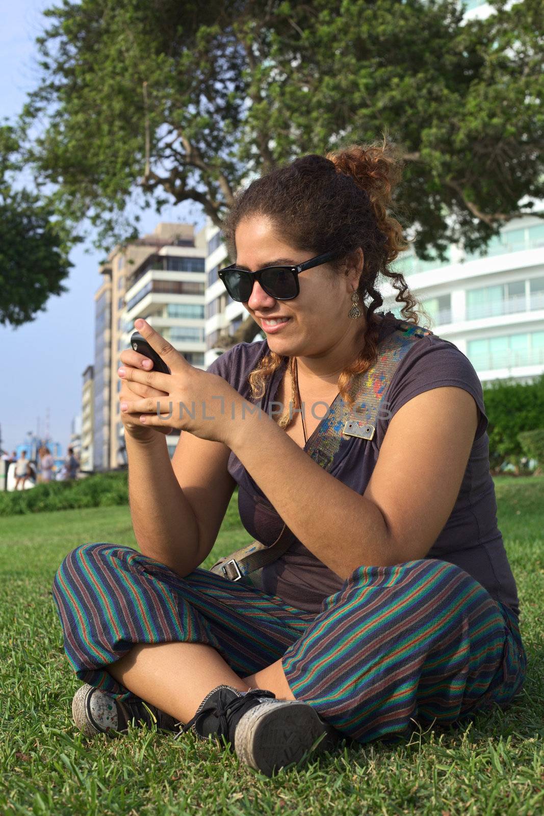 Beautiful smiling young Peruvian woman text messaging on mobile phone in park (Selective Focus, Focus on the face of the woman)