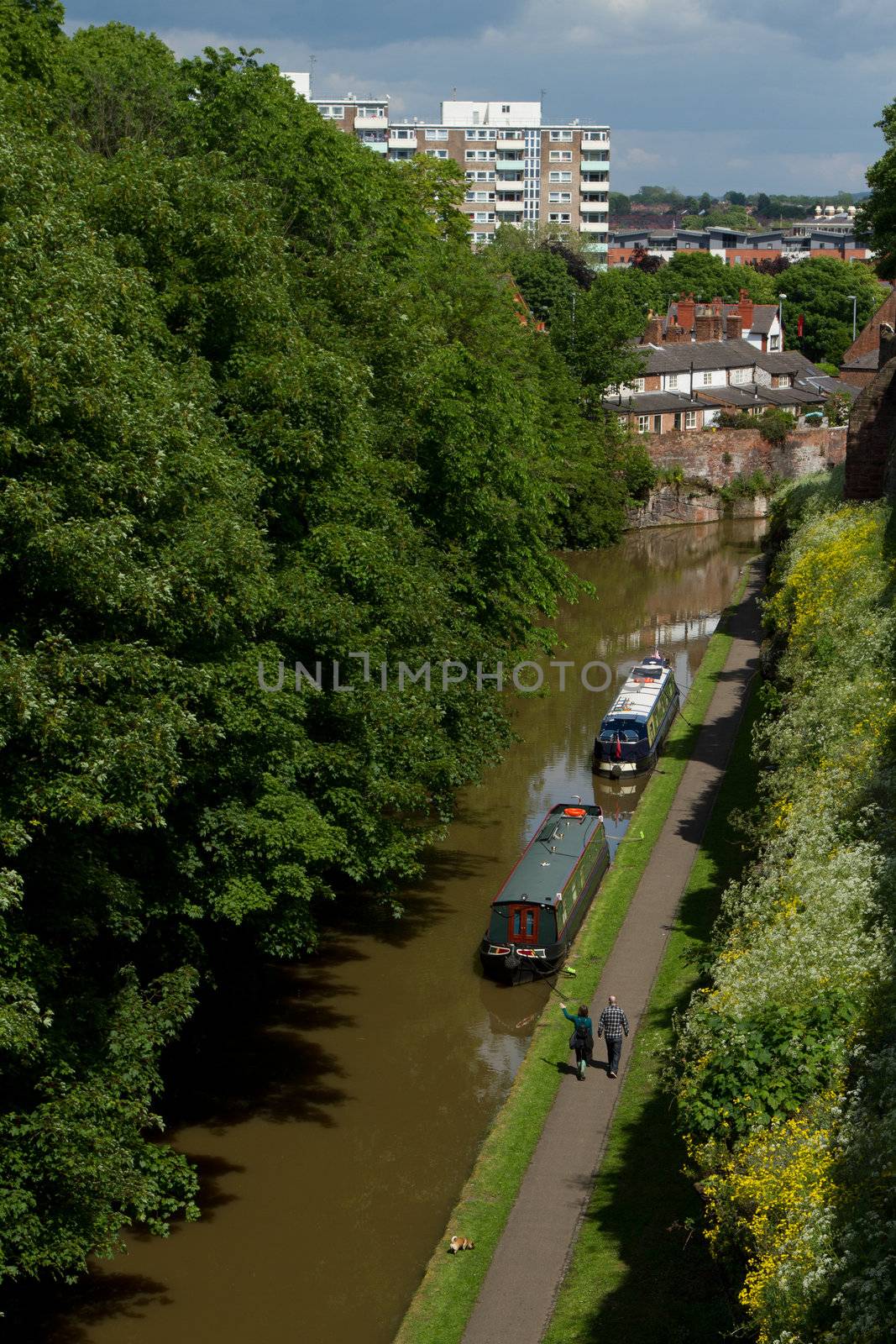 A waterway, canal, with brown water flanked by trees and two barges moored by a towpath with a couple walking on a sunny day.