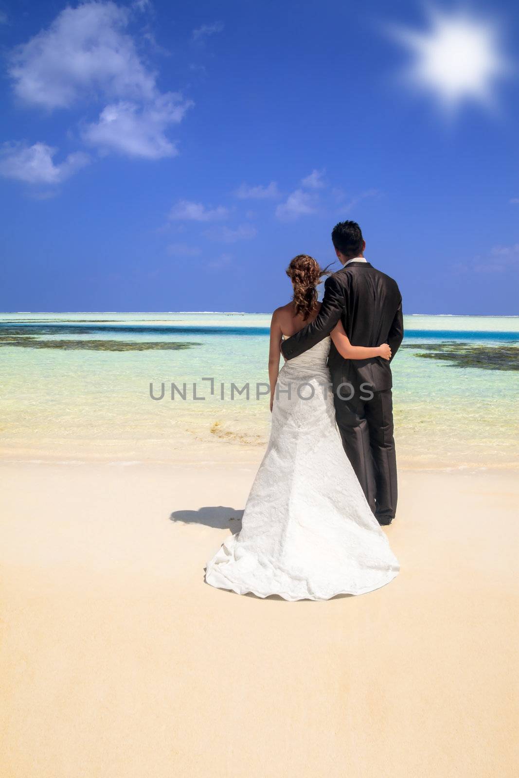 Bride and groom standing arm in arm facing away from the camera looking out over the ocean on an idyllic tropical beach