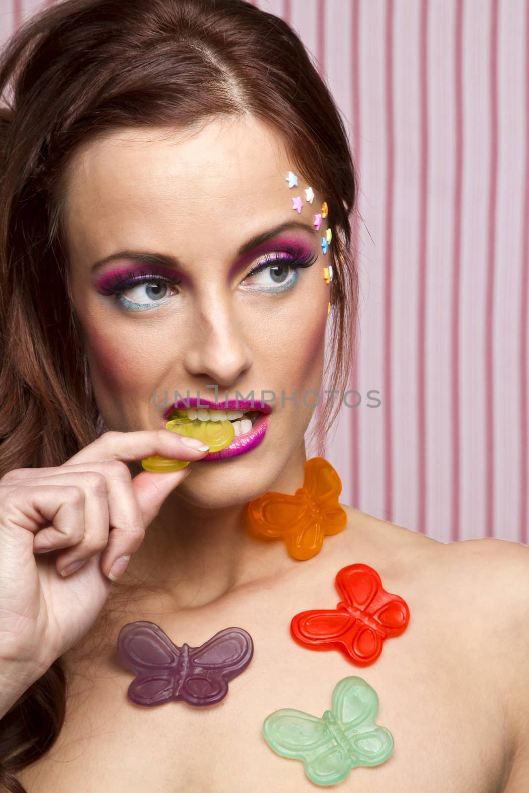 Young woman with colorful makeup and star candy glued to her face and gummy butterfly glued to her body, sucking on the wing of a yellow butterfly