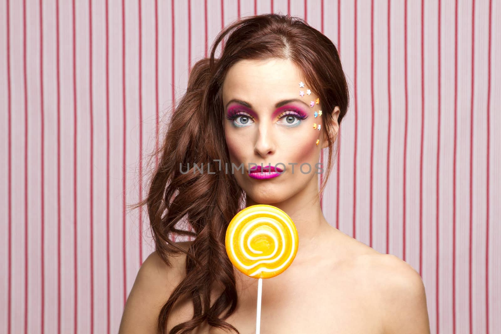 Young woman with colorful makeup and star candy glued to her face, holding an orange lollipop