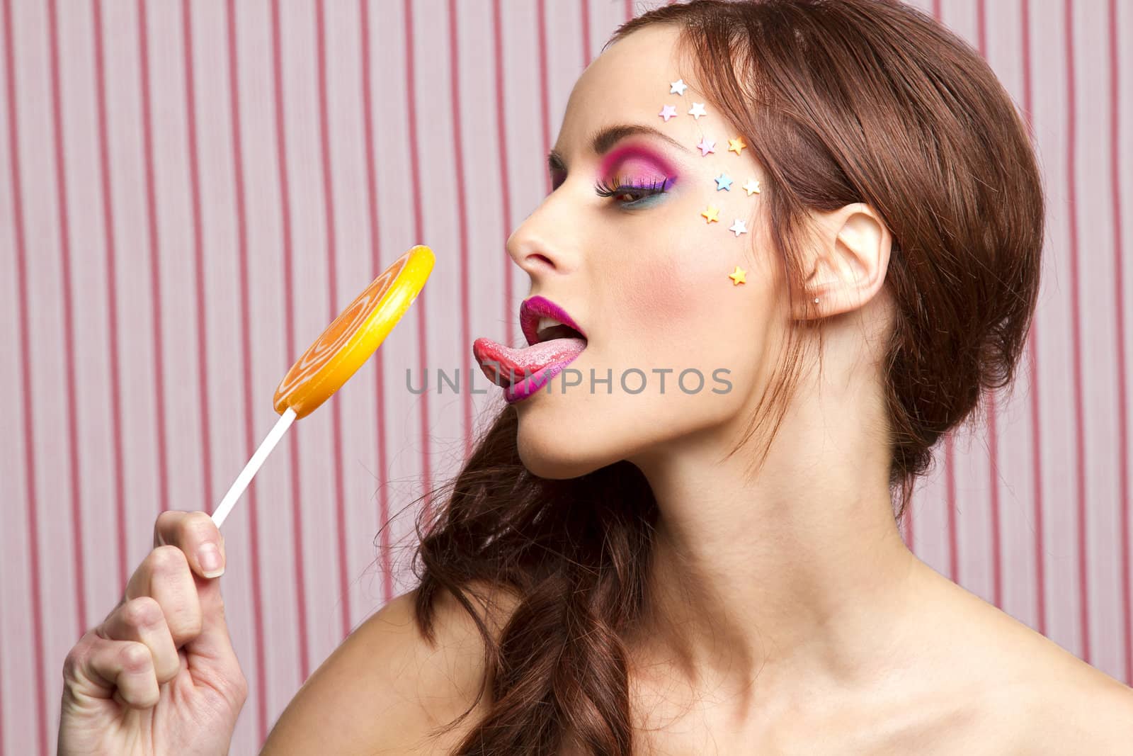 Young woman with colorful makeup and star candy glued to her face, licking an orange lollipop