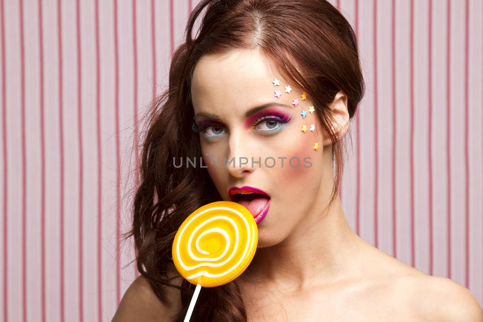 Young woman with colorful makeup and star candy glued to her face, licking an orange lollipop