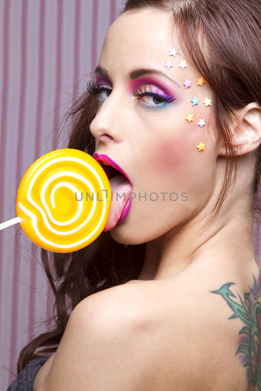 Young woman with colorful makeup and star candy glued to her face, licking an orange lollipop
