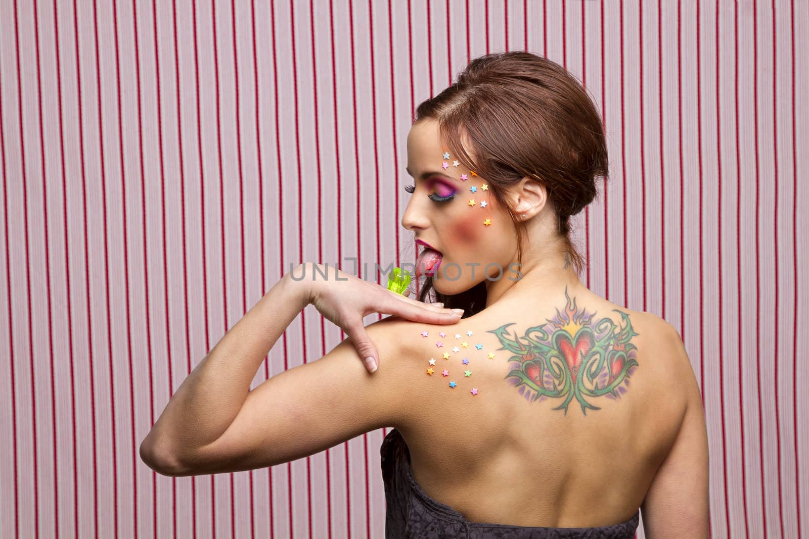 Young woman with colorful makeup and star candy glued to her face and body, licking a lollipop ring