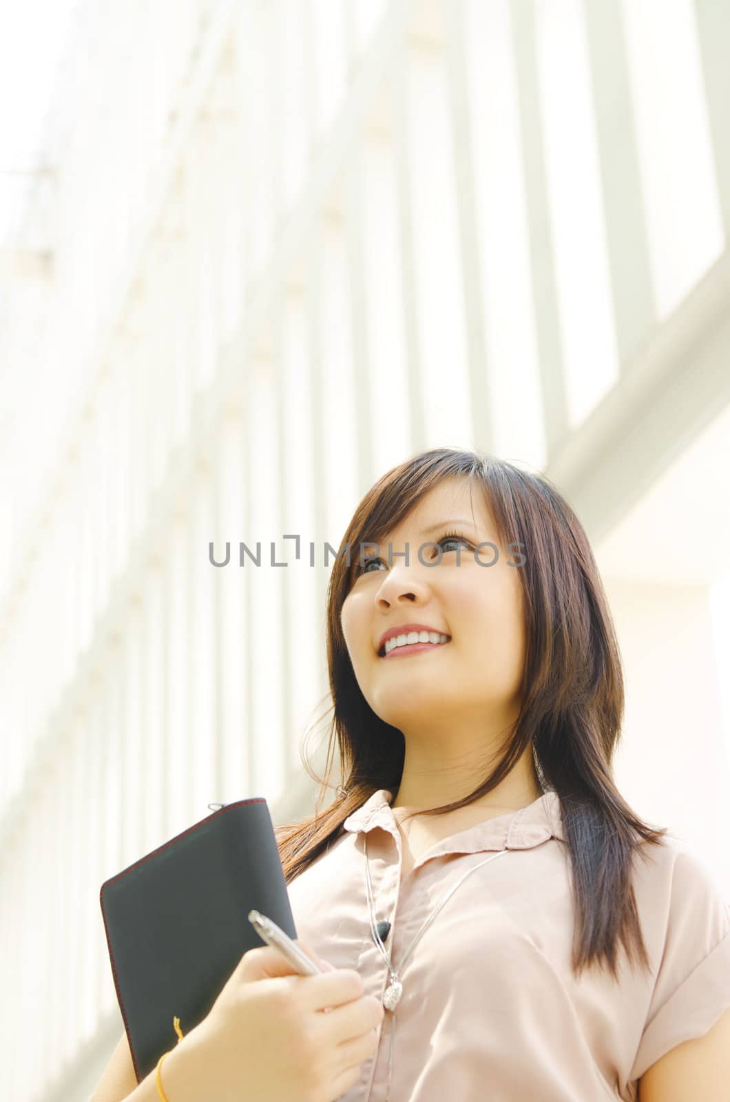 A young Asian woman looking far away to bright light in front of a modern office building, with diary on hand.