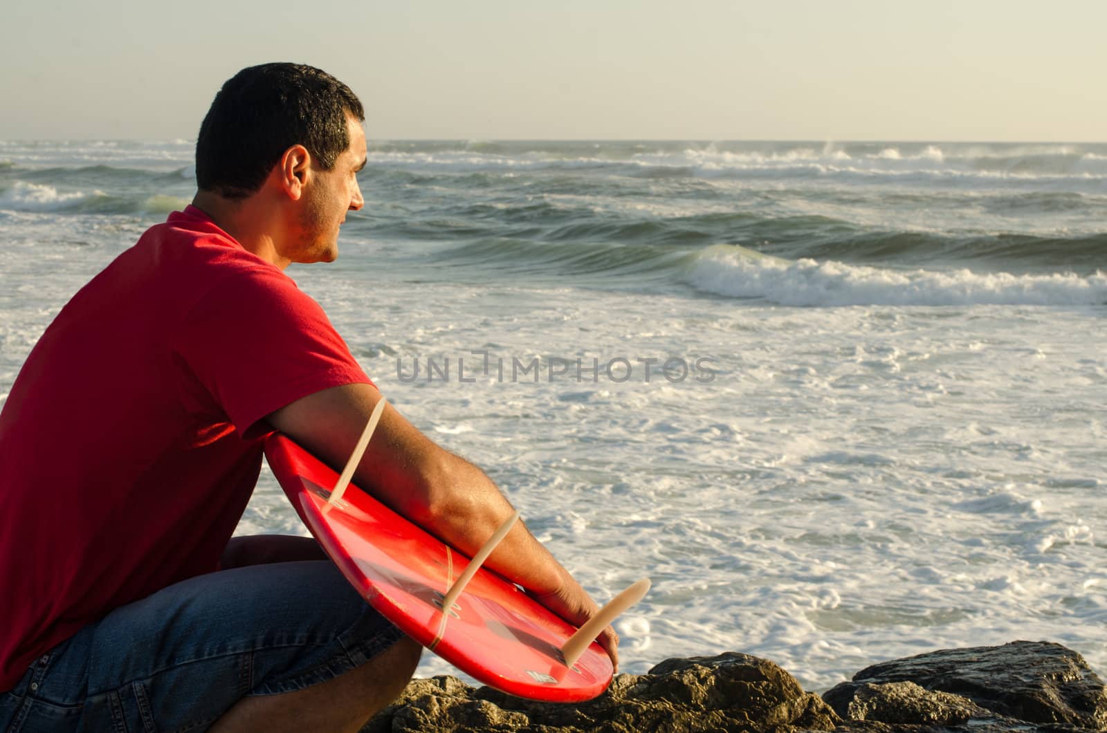A surfer watching the waves sitting down with his arms around his surfboard.