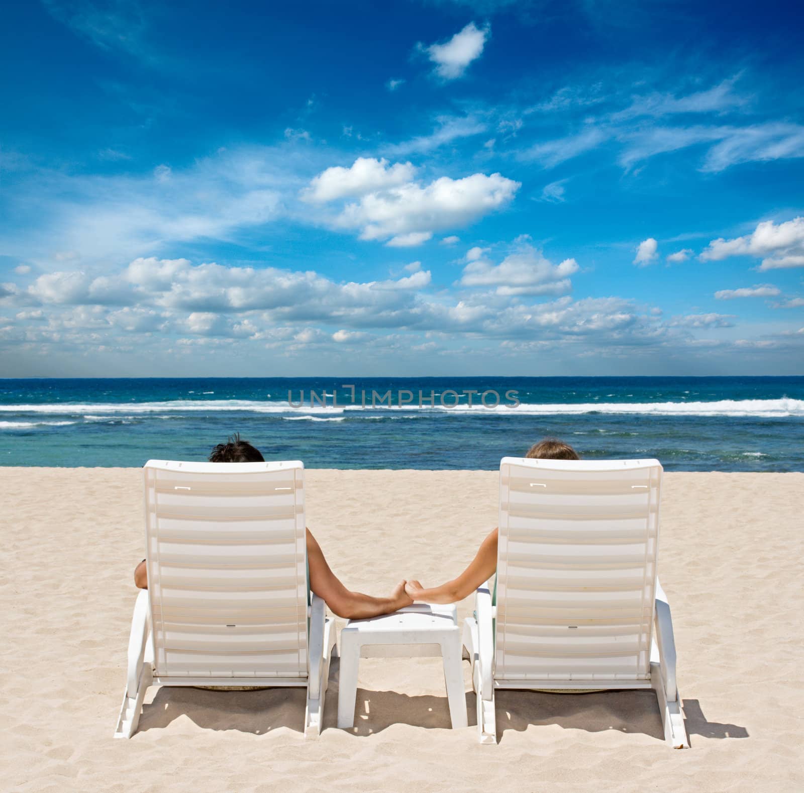 Couple in beach chairs holding hands near ocean by dimol