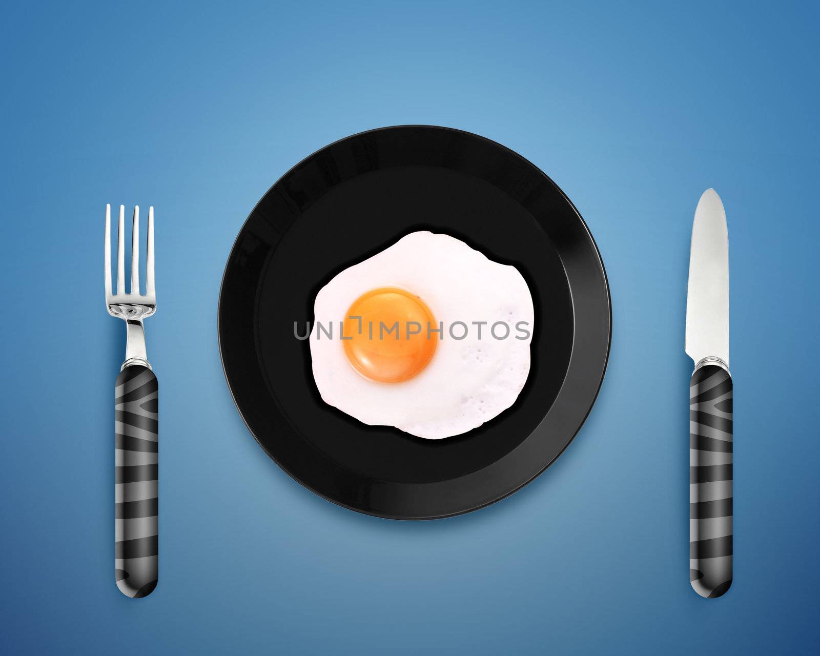 top view of fried egg on black Plate between silver knife and fork on orange background.