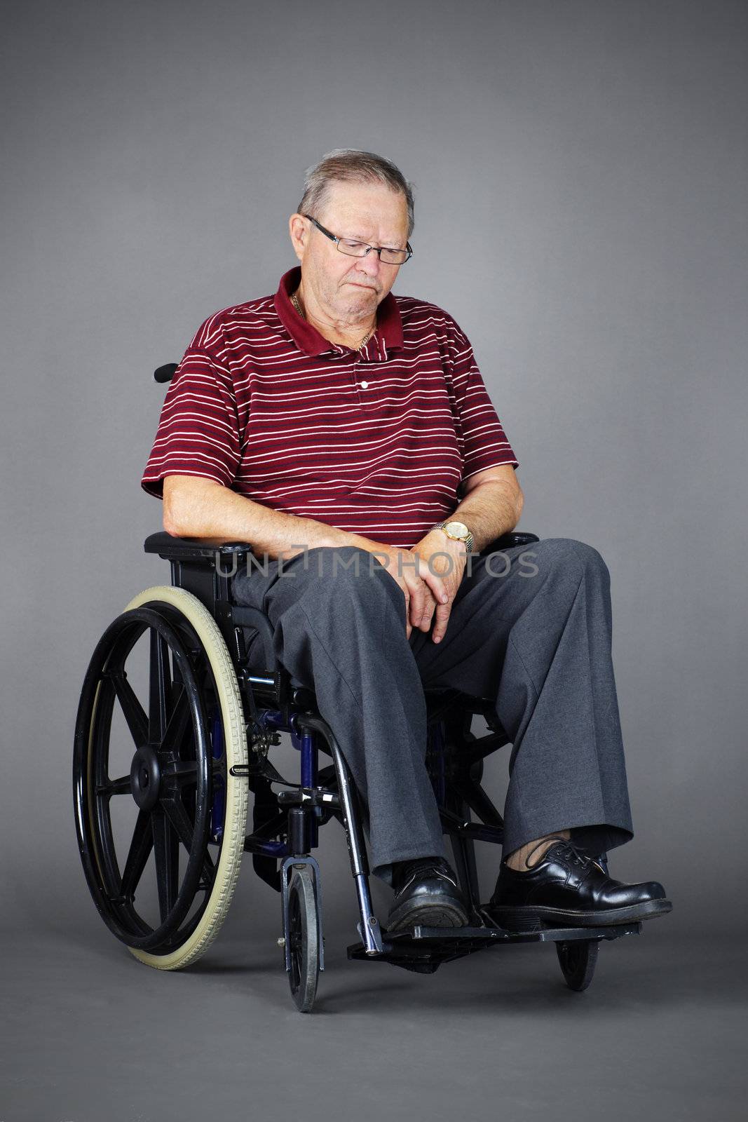 Sad or depressed senior man in a wheelchair, looking down, studio shot over grey background.
