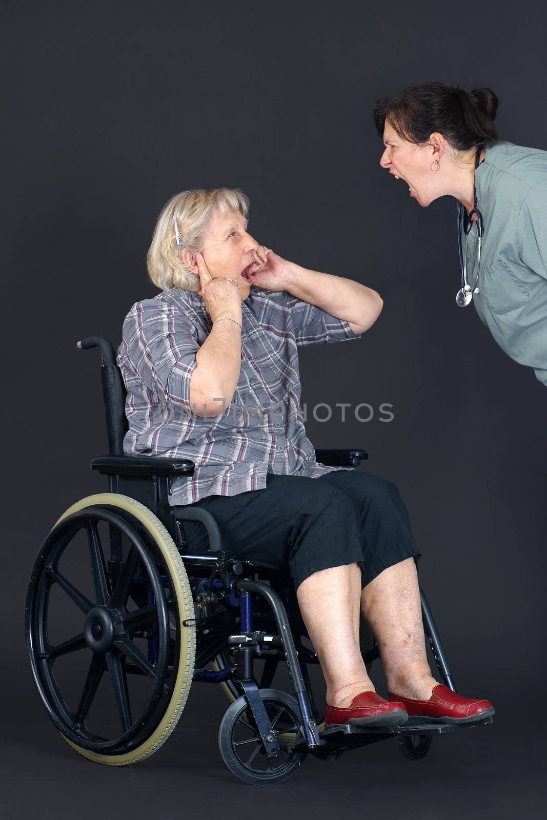 Elder abuse concept with a senior woman in a wheelchair crying and covering her ears as a middle age nurse or other health care worker is yelling at her.