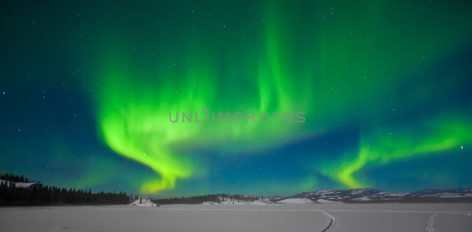 Northern Lights (Aurora borealis) over moon lit snowscape of frozen lake and forested hills.