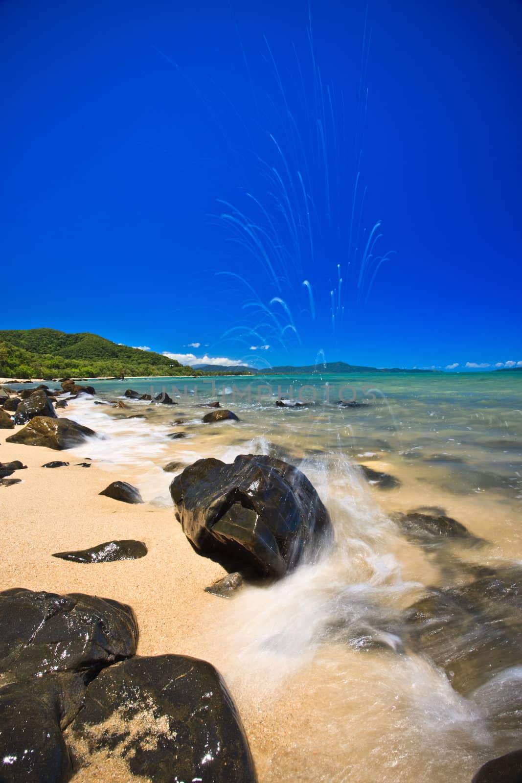 Water breaking over a rock at the seaside on a beautiful sandy beach scattered with rocks alongside an azure blue tropical ocean