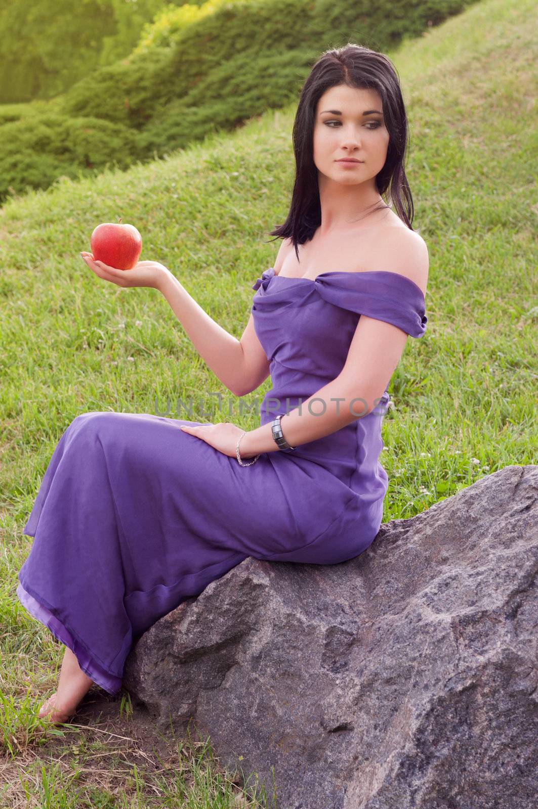 Young girl in evening dress sitting on rock and holds apple in his hand.
