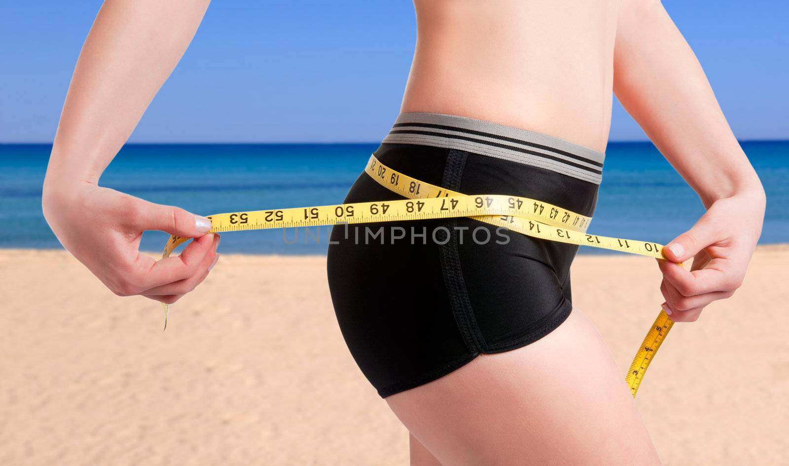 Woman measuring her waist with a yellow measuring tape. Beach background.