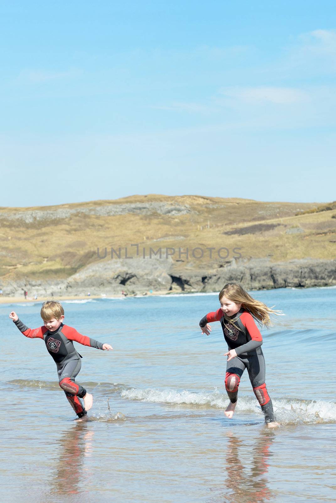 brother ans sister running in ad out of the sea in wetsuits having fun on holiday