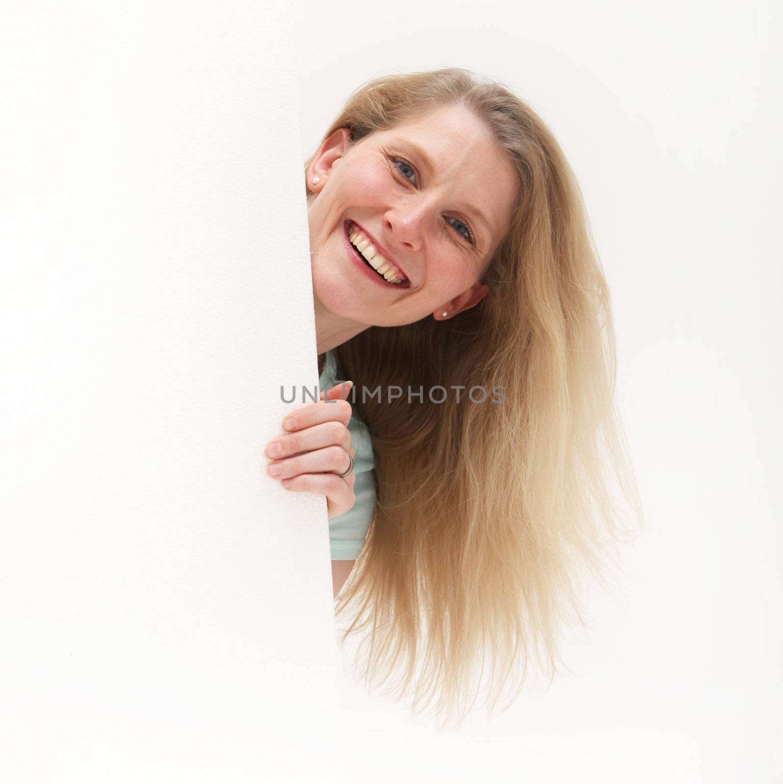 Vivacious woman popping her head around the side of a blank poster or signboard and smiling in endorsement of the product or advertising