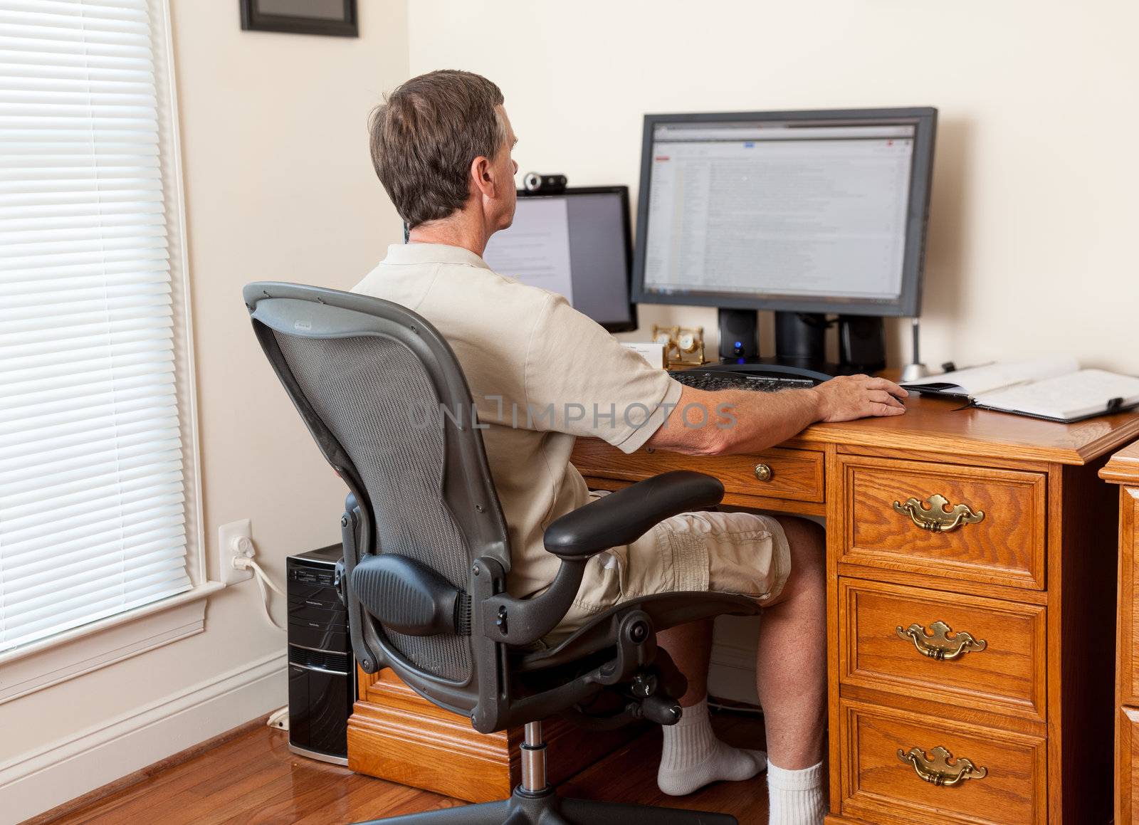 Senior caucasian man working from home in shorts with desk with two monitors