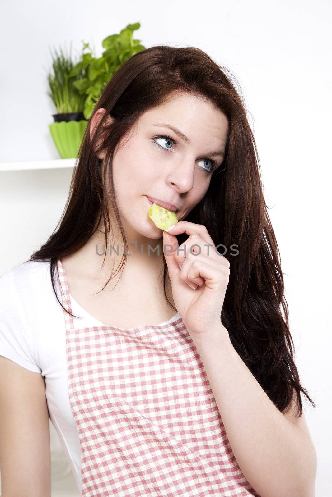 young woman in kitchen nibbling on a cucumber looking to side