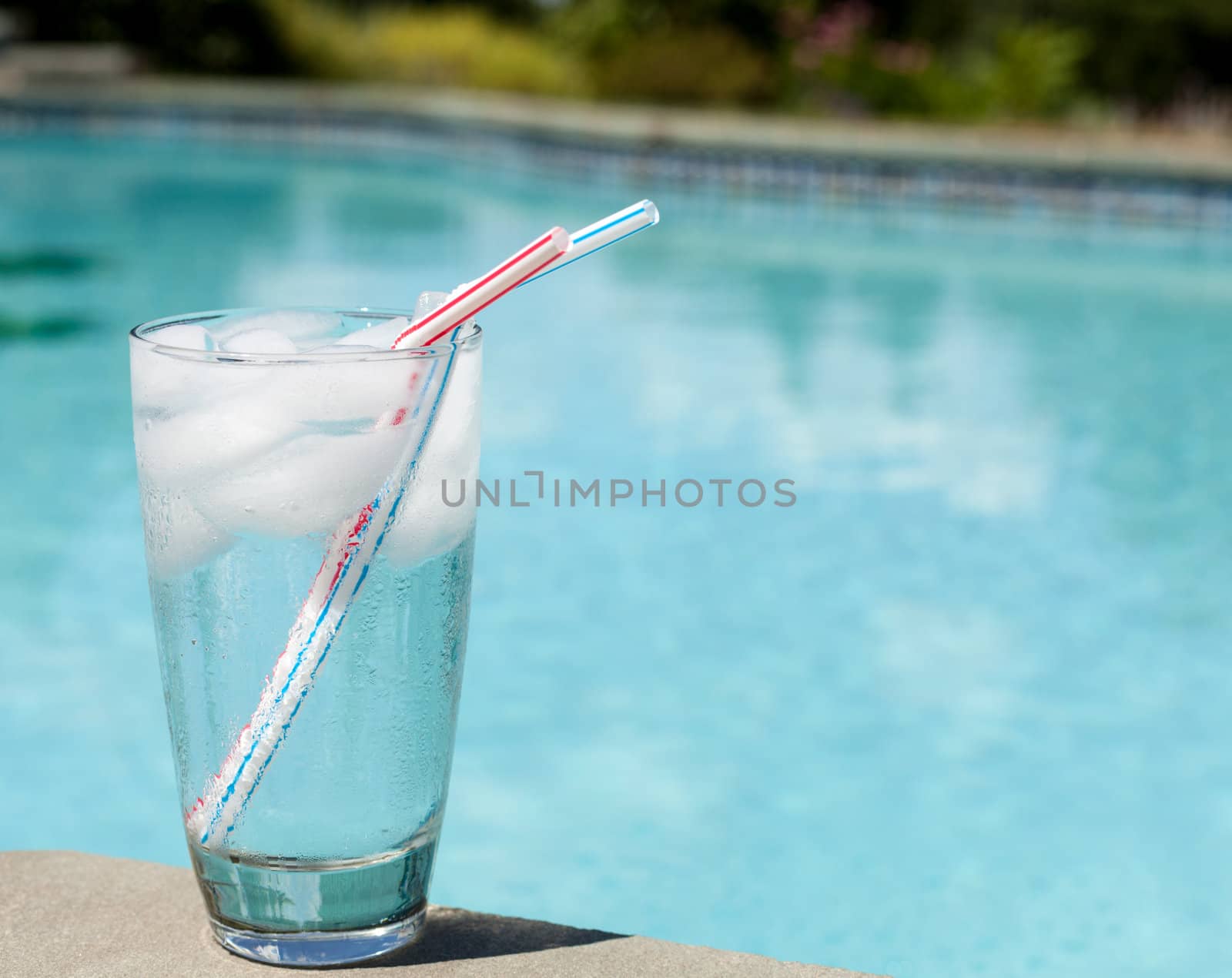 Plain glass of water with ice cubes on side of blue swimming pool