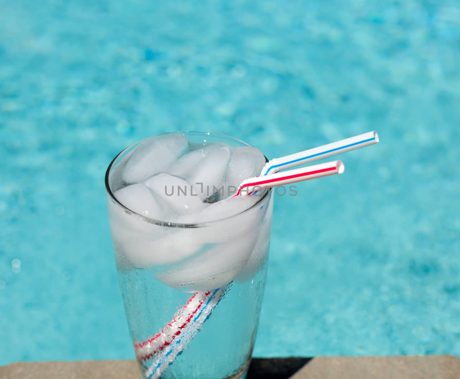 Plain glass of water with ice cubes on side of blue swimming pool