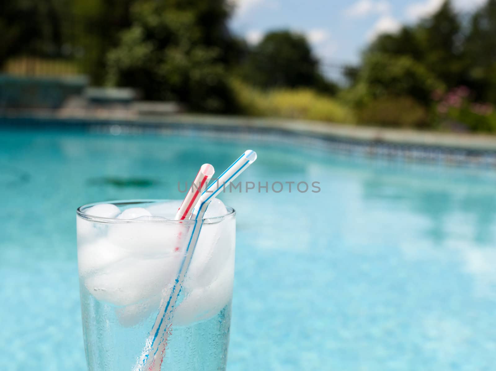 Plain glass of water with ice cubes on side of blue swimming pool