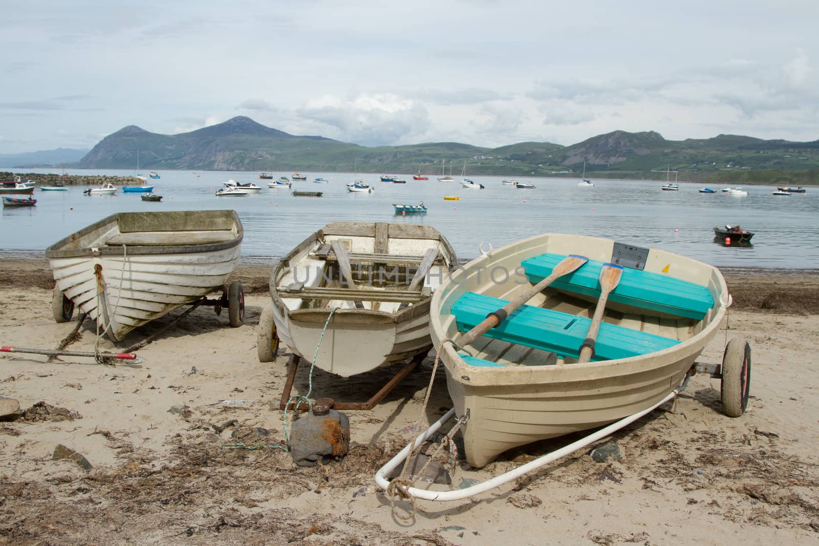 Wooden rowing boats on hand pulling trailers on a sandy beach with the sea, boats and a mountain in the distance.