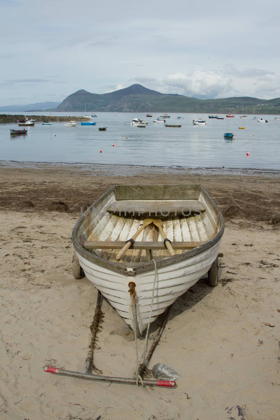 A wooden, white painted, rowing boat on a hand pulling trailer on a sandy beach with the sea, boats and a mountain in the distance.