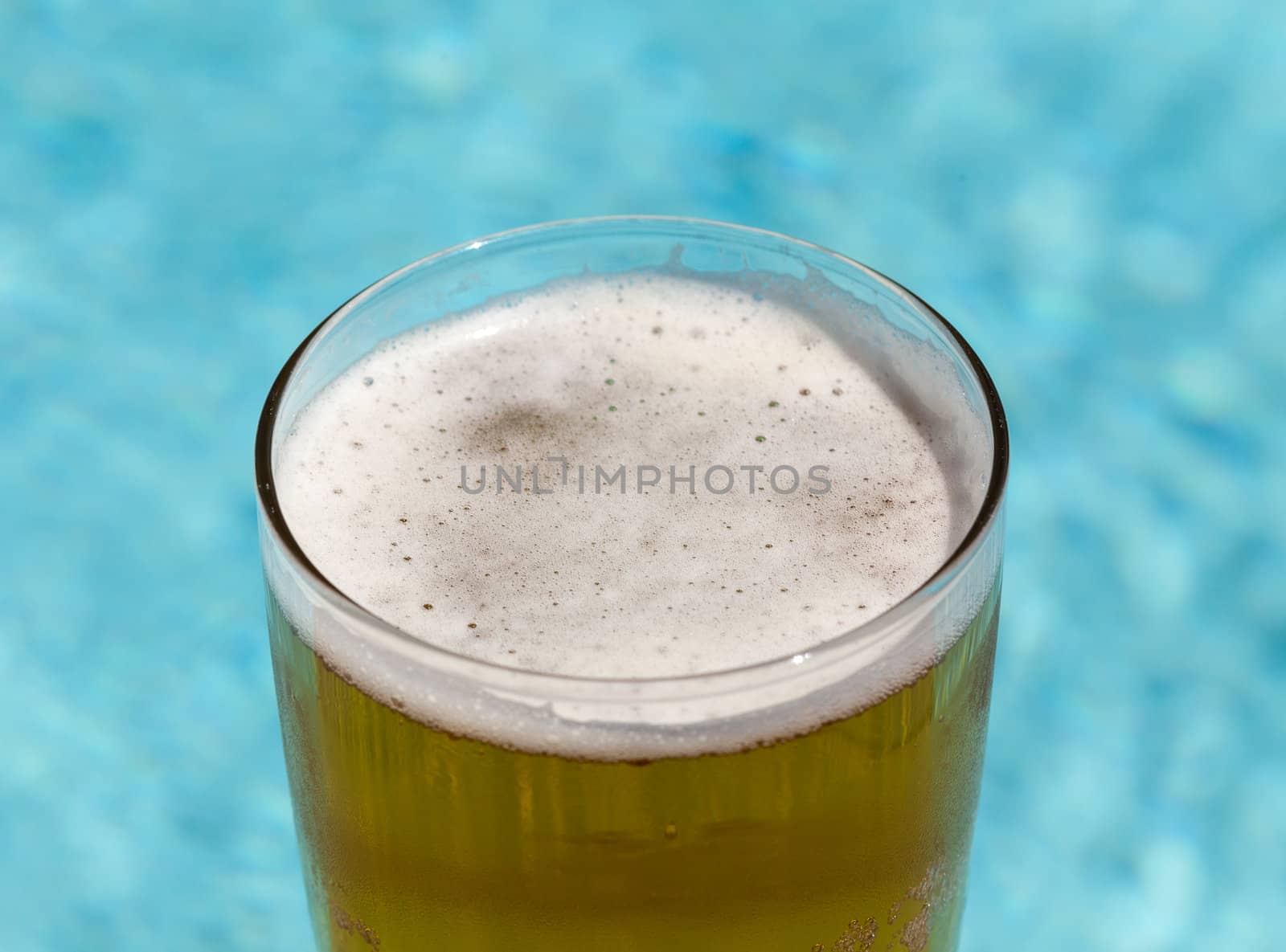 Plain pint glass of cold lager beer sitting on edge of blue swimming pool