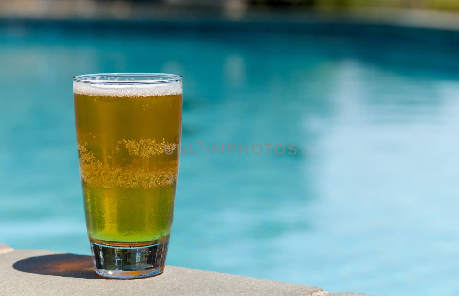 Plain pint glass of cold lager beer sitting on edge of blue swimming pool
