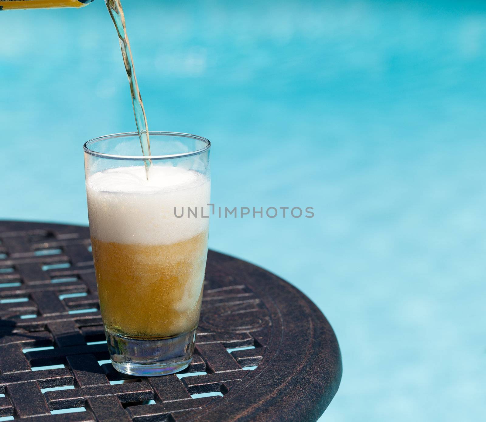 Plain pint glass of beer being poured sitting on table by blue swimming pool