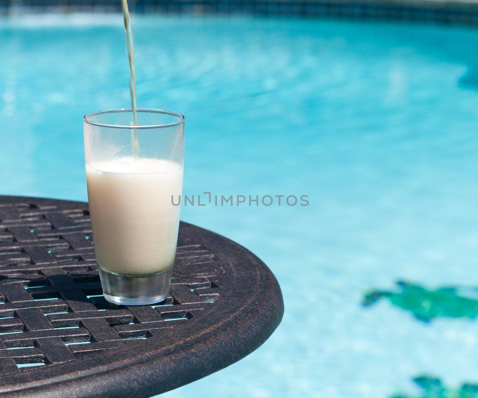 Plain pint glass of beer being poured sitting on table by blue swimming pool