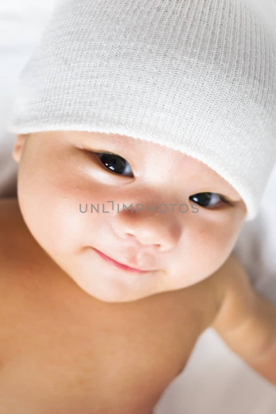 A cute little baby is looking into the camera is wearing a white cap smiling 