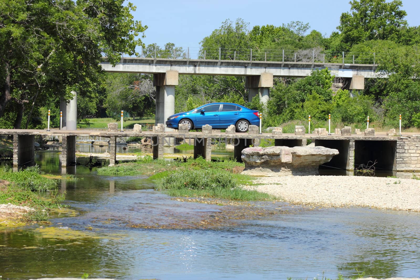 Blue car driving over an old small bridge with freeway in background.