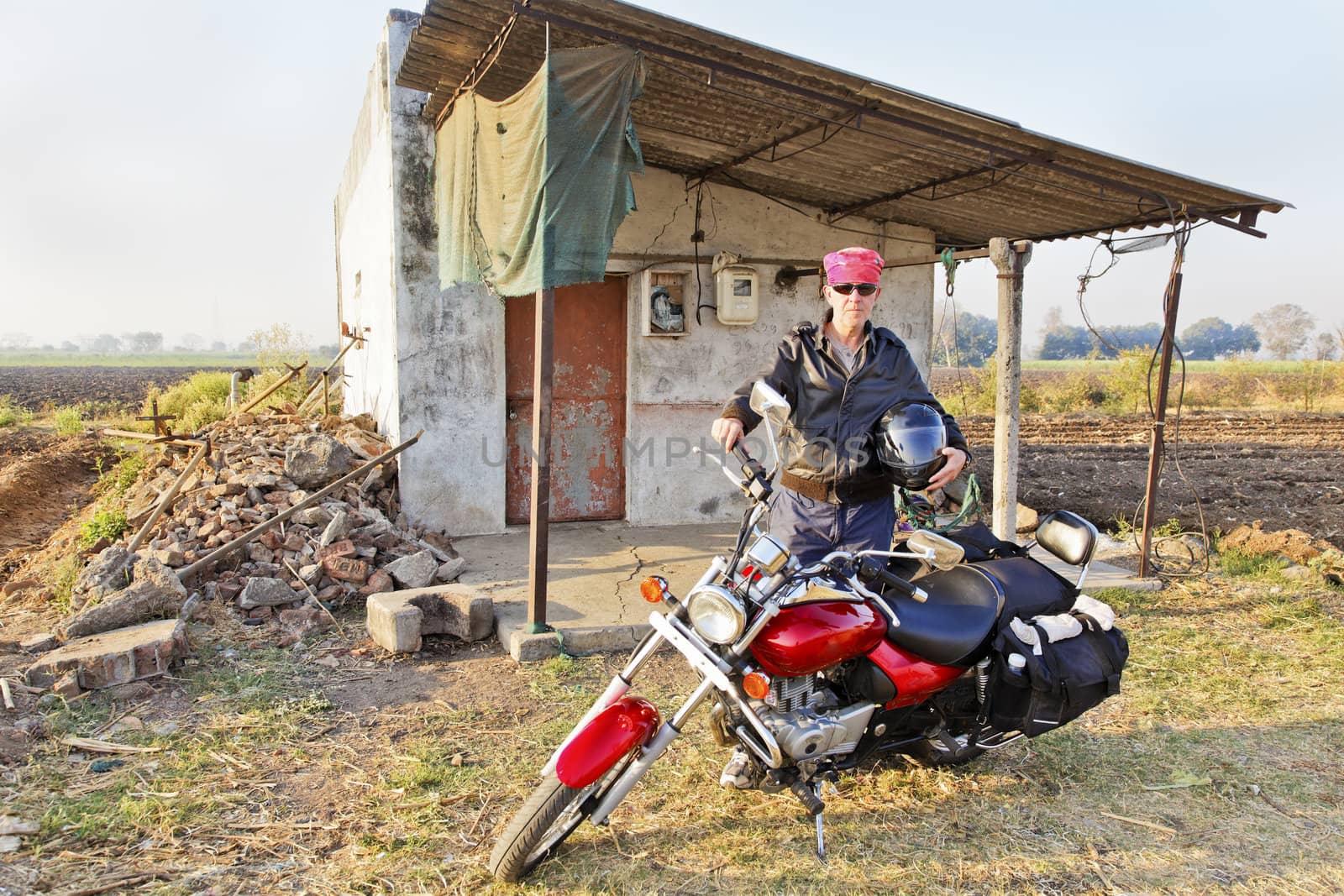 Caucasian motorcycle ride holding a helmet parked outside a rural hut earing a red bandanna