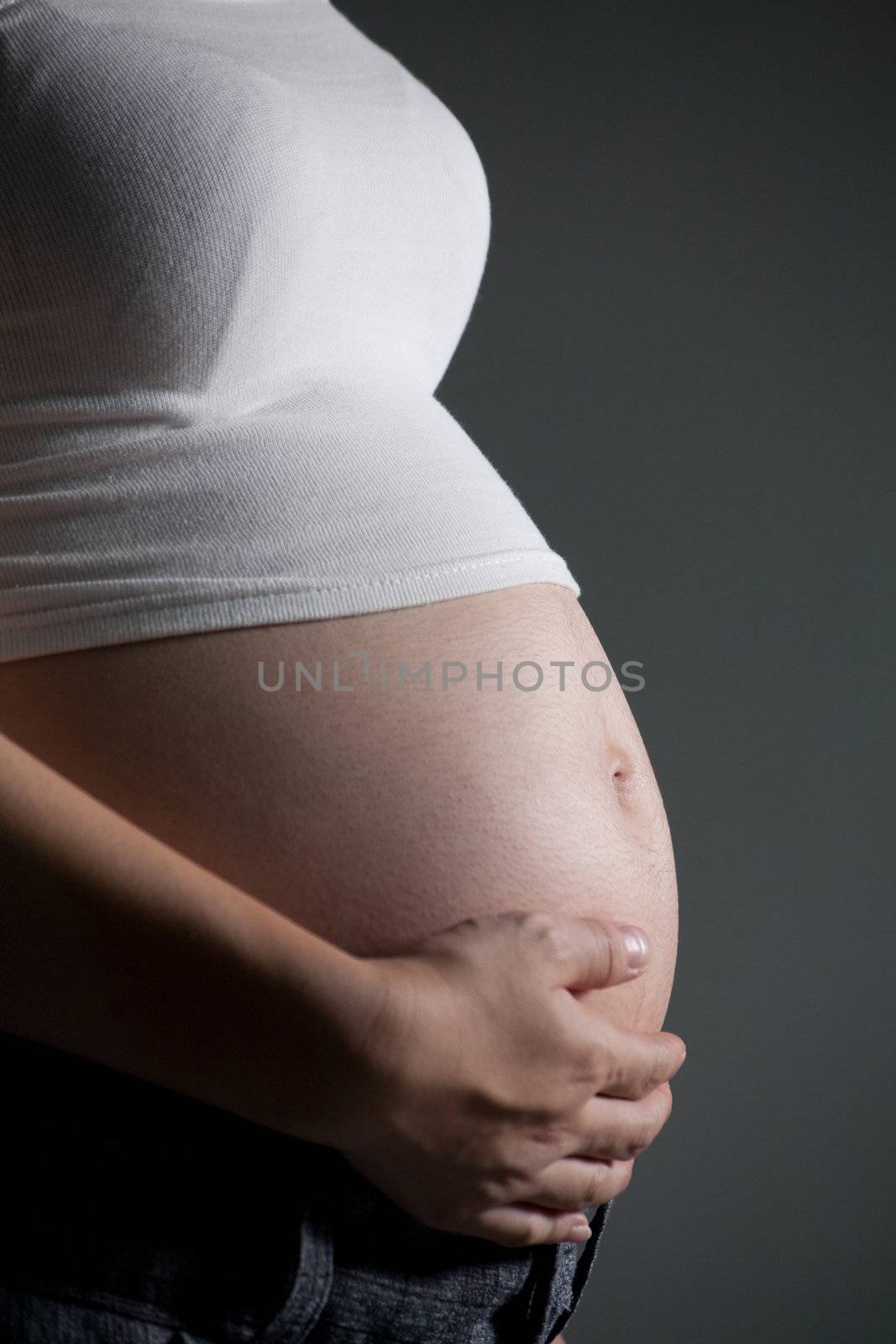 Close-up of a pregnant woman caressing her belly while standing against a gray background.