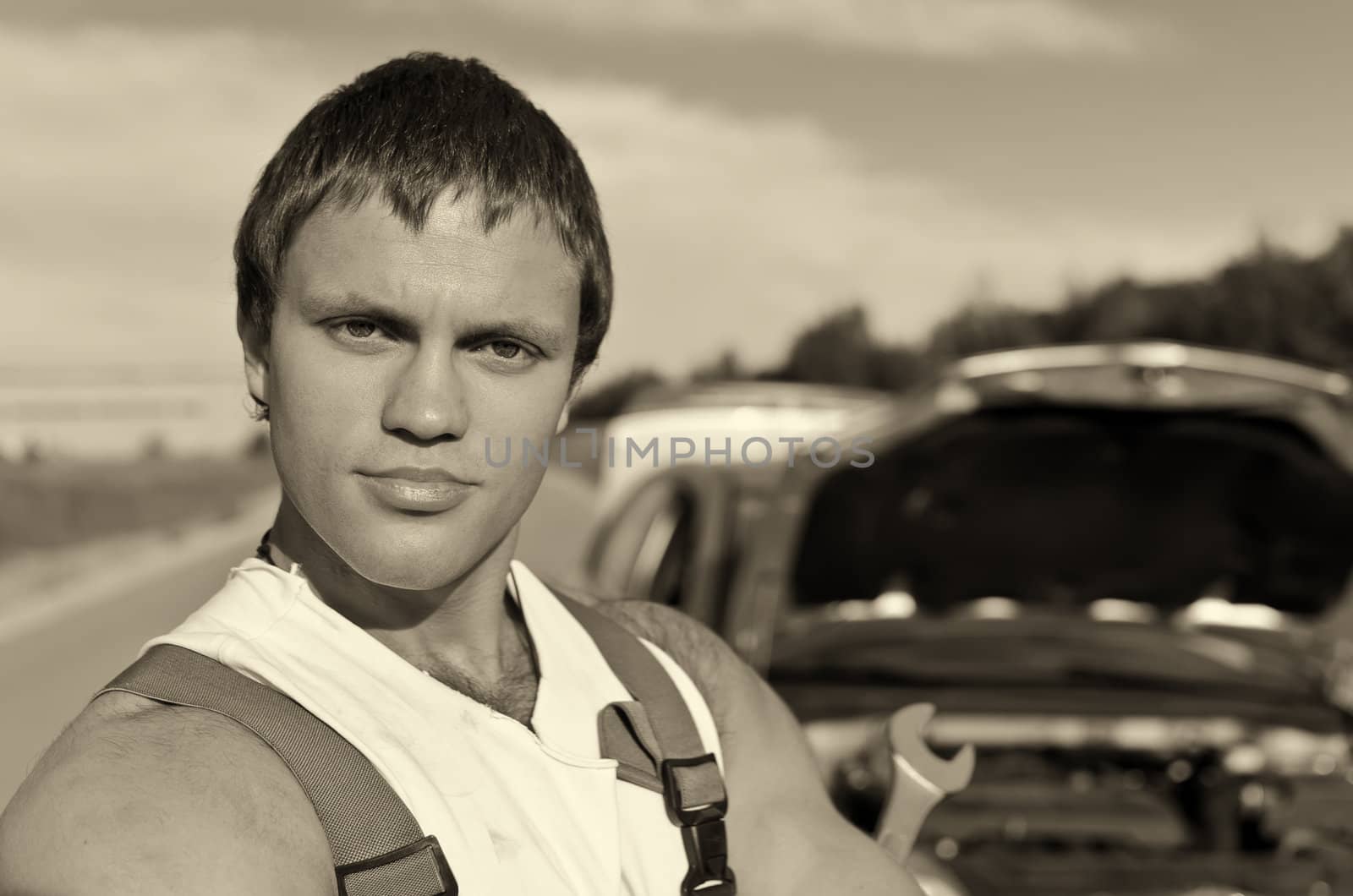 Black and white portrait of a hadsome mechanic with a broken car on background